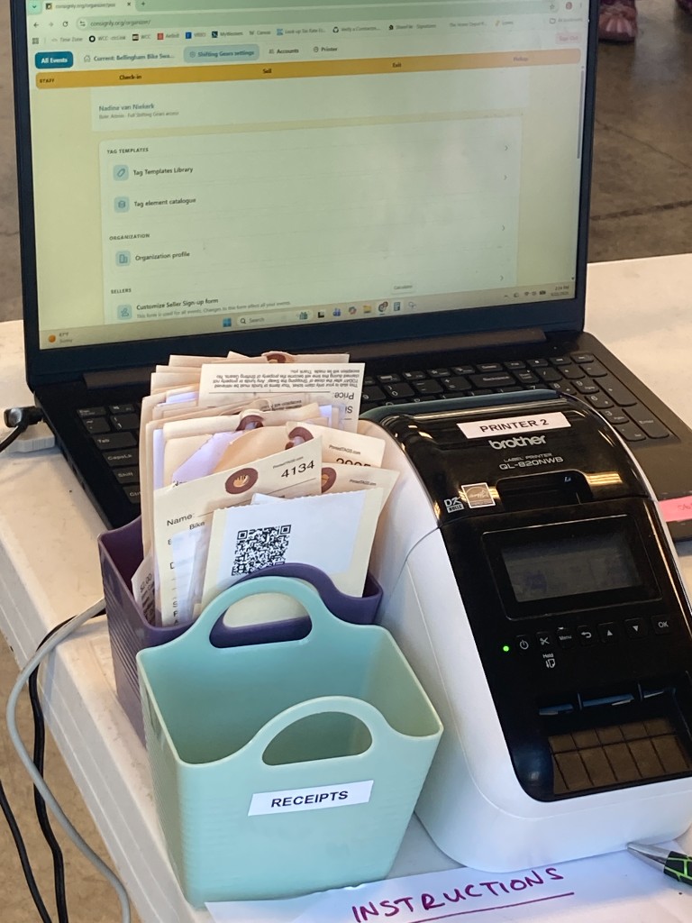 Consignly on a laptop beside a Brother label printer with bins of printed tags and receipts